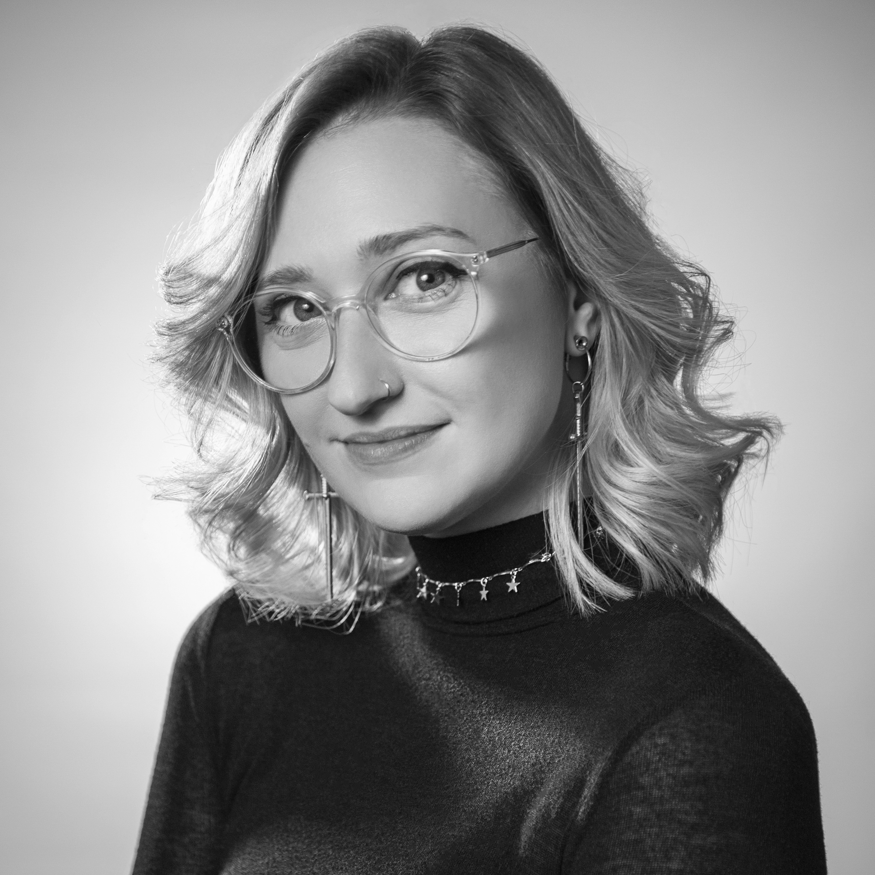 A black and white headshot of Lindsey. She is wearing glasses and has shoulder length wavy hair.